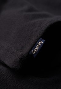 Camiseta de algodón negra con un primer plano de su textura suave y una etiqueta azul marino cosida con "Superdry". Presenta costuras limpias.