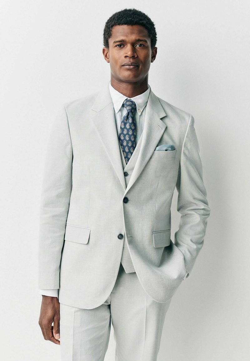 Homme portant un costume gris clair avec une chemise blanche et une cravate bleue à motifs, debout avec une main dans la poche sur un fond blanc.