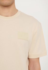 Beige pamut póló kerek nyakkivágással, amelyen a bal mellrészen egy texturált, domború EA7 logófolt található. Rövid ujjú, sima textúrájú.