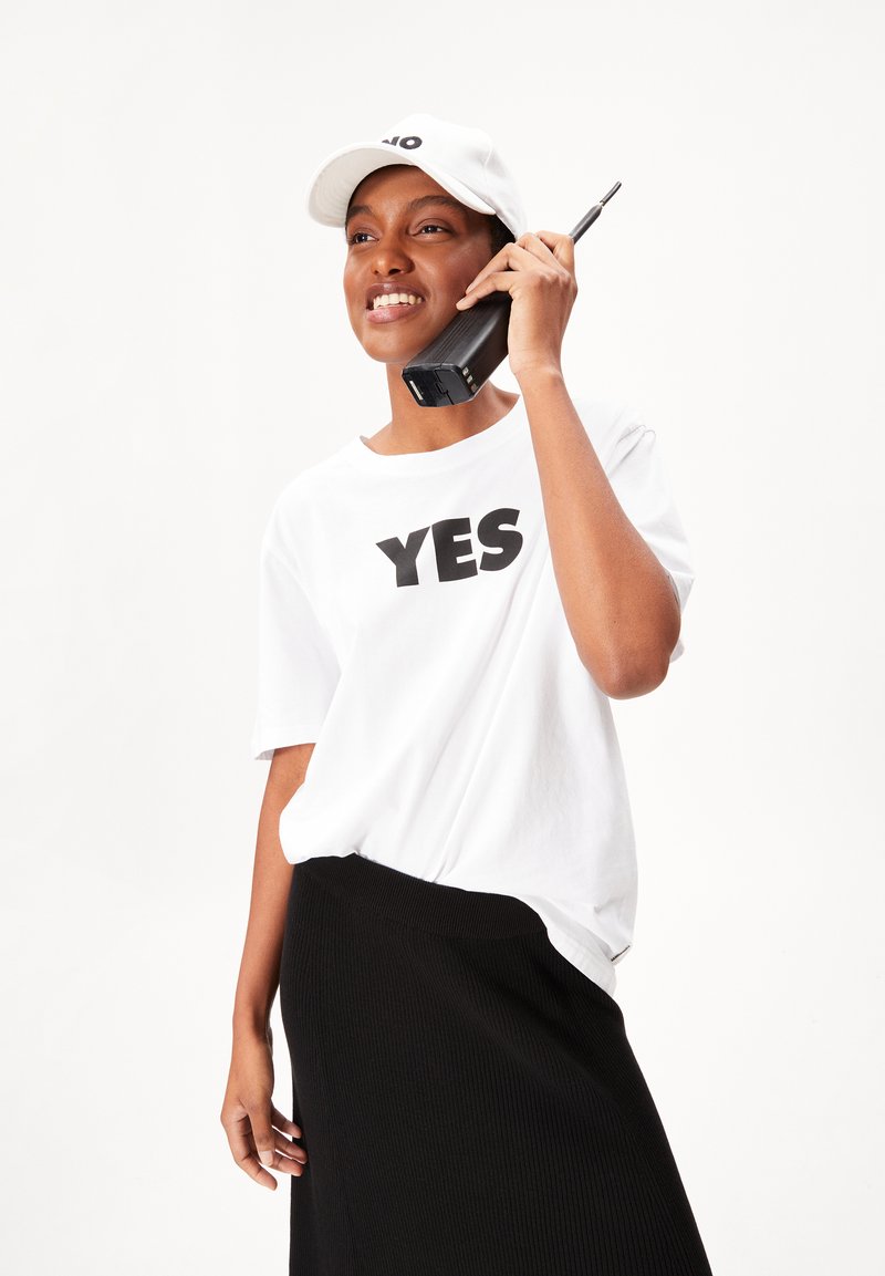 T-shirt blanche avec un texte noir en gras "OUI", associée à une jupe noire à côtes. Tient un téléphone mobile vintage noir, portant une casquette blanche.