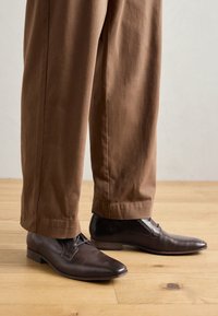 Chaussures en cuir marron texturé avec un bout pointu et de subtils perforations, assorties à un pantalon marron ample, sur un sol en bois.