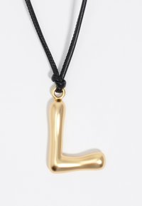 Pendente in metallo dorato a forma della lettera "L," attaccato a una corda di pelle nera con un nodo e un semplice anello dorato.