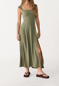 Vestido midi verde con escote cuadrado, hombros fruncidos, abertura lateral y dobladillo fluido. Combinado con sandalias marrones oscuras. Textura de tela suave.