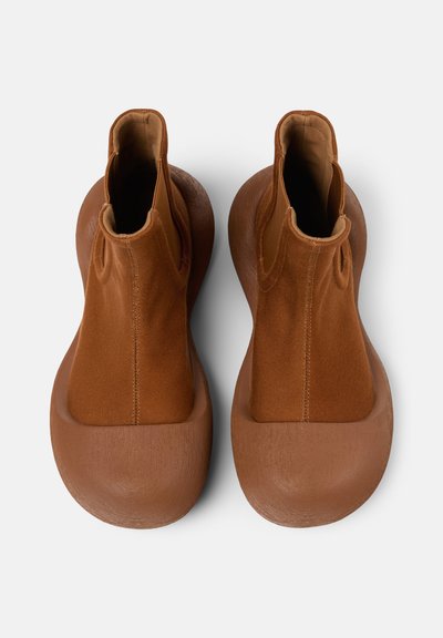 Bottes chevilles brunes avec une semelle en caoutchouc texturée et un dessus en tissu lisse. Panneaux latéraux élastiques pour un enfilage facile. Design à bout arrondi.
