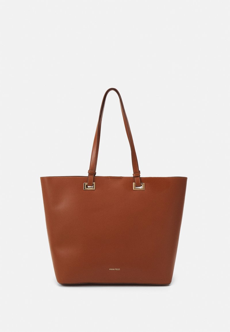 Sac fourre-tout en cuir marron avec deux longues poignées et une garniture carrée dorée sur un fond blanc uni.