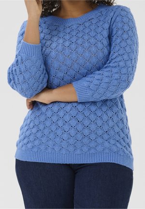 Femme en pull en maille bleu texturé à manches longues et jean bleu foncé, bras croisés, sur fond clair uni.