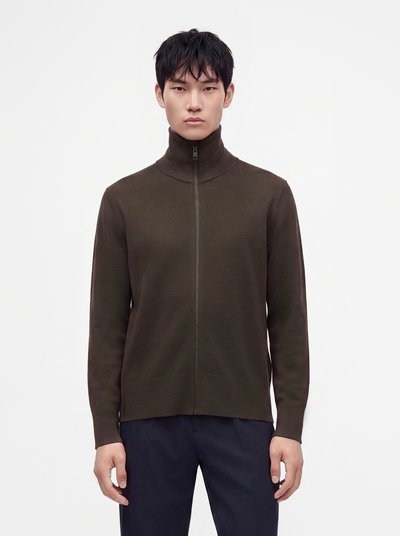 Jeune homme portant un pull zippé marron foncé avec un col montant et un pantalon bleu marine, debout devant un fond blanc uni.