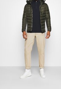 Doudoune vert olive avec capuche et fermeture éclair, portée sur un haut sombre ; associée à un pantalon beige et des baskets blanches.