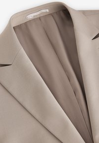 Blazer beige clair avec col à revers cranté, doublure intérieure lisse et petite étiquette en tissu cousue à l'intérieur de l'encolure.