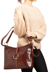 Braune Lederhandtasche mit glänzender Oberfläche, abgerundetem Metallakzent und verstellbarem Tragegurt, gehalten von einer Person, die einen strukturierten beigen Pullover trägt.