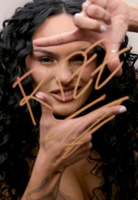 Person, die Handgesten vor dem Gesicht macht, mit lockigem schwarzen Haar. Hände mit Ringen verziert; zeigt handgeschriebene Texte in Braun auf einer Oberfläche.