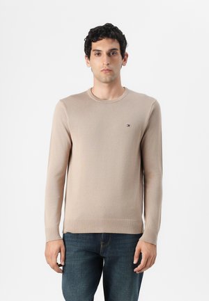 Suéter de punto beige claro con cuello redondo y mangas largas. Presenta un pequeño logo en el pecho y un dobladillo y puños acanalados. Combinado con jeans oscuros.