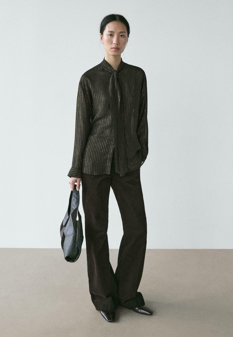 Metaliczna czarna koszula na guziki w paski z kołnierzykiem wiązanym na kokardę, zestawiona z szerokimi ciemnobrązowymi spodniami i czarną torbą na ramię. Czarne, spiczaste buty.