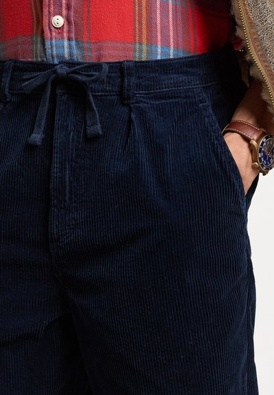 Mörkblå kordflosspants med dragsko i midjan, sidofickor och en ribbad textur, kombinerade med en röd rutig skjorta.