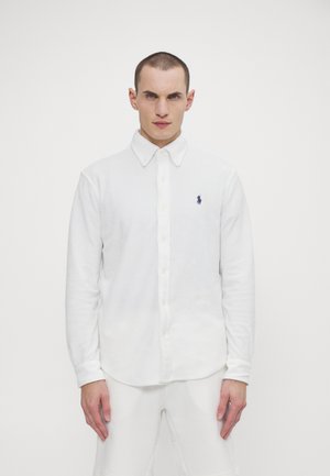 Camisa blanca de manga larga con botones, hecha de una tela suave, que presenta un cuello clásico y un pequeño logo en azul marino en el lado izquierdo del pecho.
