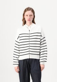 Hvit strikket cardigan med svarte horisontale striper, glidelås foran, ribbestrikket tekstur, og oversized passform; brukt over en skjorte med krage.
