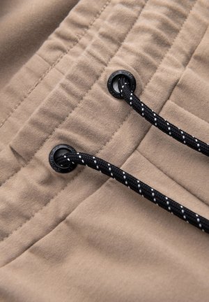Beige stof met een gladde textuur; beschikt over zwarte koordjes met witte accenten en plastic hardware. Ontworpen voor een verstelbare pasvorm.