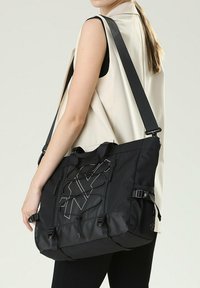 Borsa a tracolla nera in tessuto con un design geometrico, tracolla regolabile e manici superiori. Presenta tasche laterali in rete e dettagli in metallo.