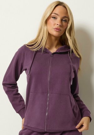 Jonge vrouw met een paarse hoodie met ritssluiting en voorzakken, staand tegen een effen lichte achtergrond.