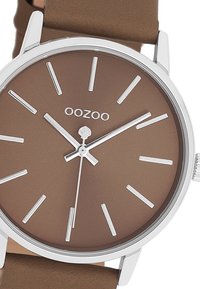 Montre marron avec des détails en argent, cadran minimaliste, longs marqueurs d'heure et bracelet marron ajustable. Logo de la marque "OOZOO" sur le cadran.