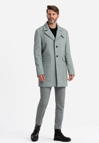 Abrigo gris de mezcla de lana con grandes solapas, botones frontales y bolsillos laterales. Llevado sobre una camisa negra y combinado con pantalones grises y botas negras.