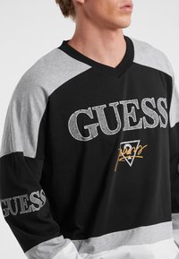 Svart och grå långärmad tröja med V-ringning, texturerad logotryck och kontrasterande färgblock på axlar och ärmar.