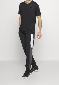 Camiseta negra con un logo blanco, pantalones de chándal en negro y gris con un panel blanco, y zapatillas negras con detalles texturizados.