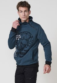 Sudadera azul marino con cuello alto, que presenta un diseño de calavera geométrico y texto. Tejido texturizado con puños y acentos negros.