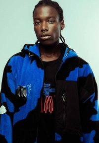 Chaqueta de forro polar azul y negra con capucha, que presenta detalles bordados y un bolsillo con cremallera. Llevada sobre una camiseta gráfica negra.