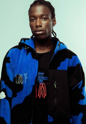 Chaqueta de forro polar azul y negra con capucha, que presenta detalles bordados y un bolsillo con cremallera. Llevada sobre una camiseta gráfica negra.