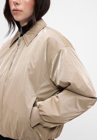 Beige, glossy bomber jacket with a zip front, large collar, and elastic cuffs. Subtle logo detail on the chest and side pockets.