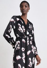 Černé dlouhé šaty s dlouhým rukávem a květinovým vzorem v bílé a červené barvě, s límečkem a páskem zdůrazněným stříbrnou sponou. Hladká textura látky.