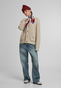 Cardigan bej cu dungi, dotat cu butoane și două buzunare frontale, purtat cu jeansi largi de culoare albastru deschis și o căciulă roșie. Eșarfa are modele geometrice colorate.