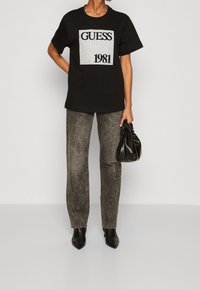 Svart bomullströja med en silverfärgad "GUESS 1981" logotyp på framsidan, kombinerad med grå högmidjade jeans och svarta spetsiga klackar.