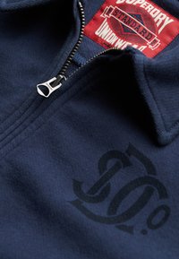 Sudadera con cremallera en azul marino, de tela texturada, con una cremallera plateada y un estampado de logo negro en la parte delantera, acompañada de una etiqueta roja.