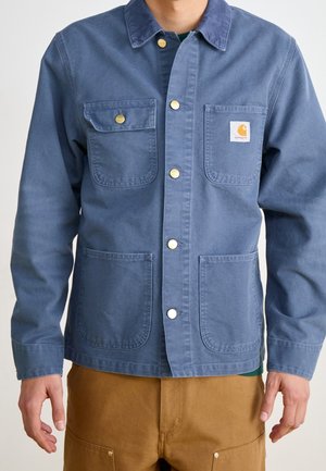 Mężczyzna ubrany w niebieską roboczą kurtkę na guziki z czterema kieszeniami, z logo Carhartt na górnej prawej kieszeni, oraz brązowe spodnie.