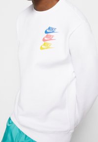 Vit sweatshirt med rund halsringning, med tre flerfärgade Nike-logotyper i blått, rosa och gult. Slät textur, avslappnad passform.