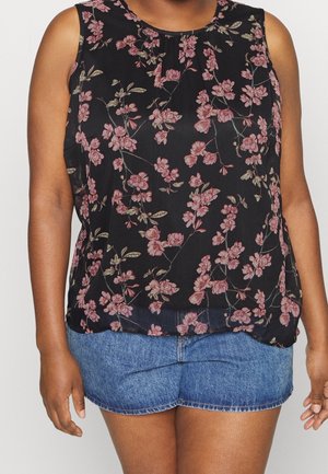 Kobieta ubrana w czarny, bezrękawnik z różowym kwiatowym wzorem i niebieskie szorty jeansowe, stoi na jednolitym tle.