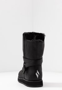 Schwarze Wildlederstiefel mit einem mit Fell gefütterten Kragen, ausgestattet mit einem verstellbaren Riemen und einer strukturierten Außensohle. Logoakzent in der Nähe der Ferse.