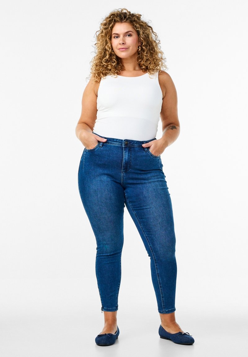 Jeans skinny taille haute bleus avec poches avant, portés avec un débardeur blanc ajusté. Le matériau semble être du denim avec une légère élasticité, texture lisse.