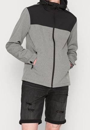 Osoba nosząca szarą i czarną kurtkę z kapturem, z rękami w kieszeniach, w połączeniu z czarnymi, podartymi szortami dżinsowymi podwiniętymi u dołu.