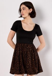 Donna con capelli corti e scuri, che indossa un vestito a maniche corte di colore nero con una gonna a stampa leopardata marrone e nera, che guarda di lato.
