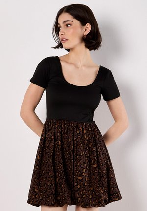 Femme aux cheveux courts et foncés portant une robe à manches courtes de couleur noire avec une jupe à imprimé léopard marron et noir, regardant sur le côté.