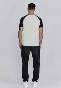 Camiseta tipo raglán en crema y negro, con mangas cortas, cuello redondo y una textura suave, combinada con jeans negros y zapatillas blancas.