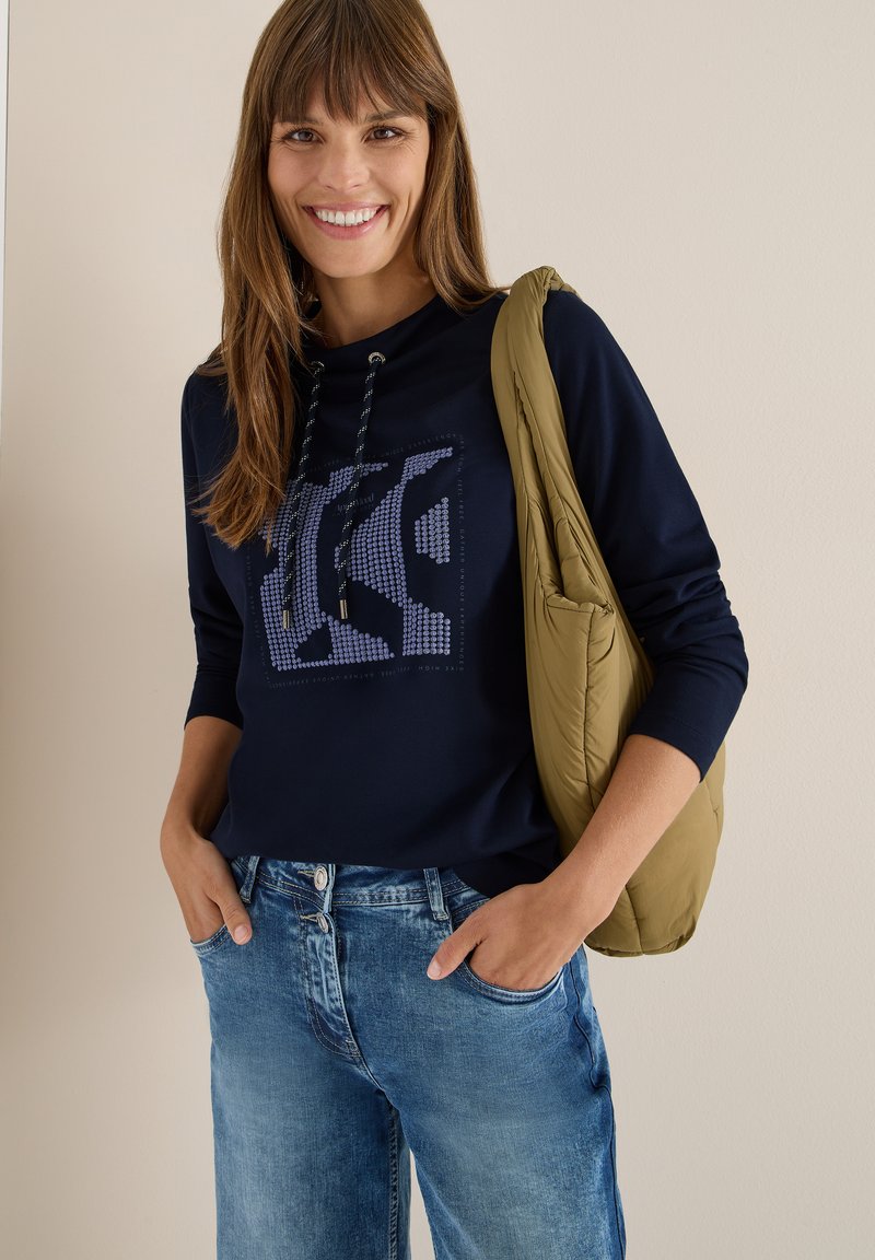 Camisa de manga larga azul marino con un diseño impreso, combinada con jeans de lavado claro. Una bolsa acolchada de color verde oliva descansa sobre un hombro.