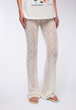 Pantaloni svasati in pizzo di colore bianco sporco con motivi a rombi. Presentano una consistenza morbida e orli arrotolati. Abbinati a calzature beige.