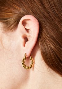 Orecchini a cerchio in oro con design a forma di cuore, caratterizzati da una finitura liscia e texture, mostrati su un orecchio con capelli marrone rossiccio.