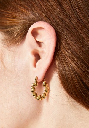 Orecchini a cerchio in oro con design a forma di cuore, caratterizzati da una finitura liscia e texture, mostrati su un orecchio con capelli marrone rossiccio.