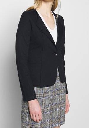 Black fitted blazer with a single button closure, notched lapels, and sleek design, paired with a checkered skirt for contrast.