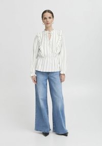 Blusa bianca a righe con colletto a volant, apertura a chiave, abbinata a jeans blu larghi e décolleté neri a punta. Tessuto dalla consistenza liscia.